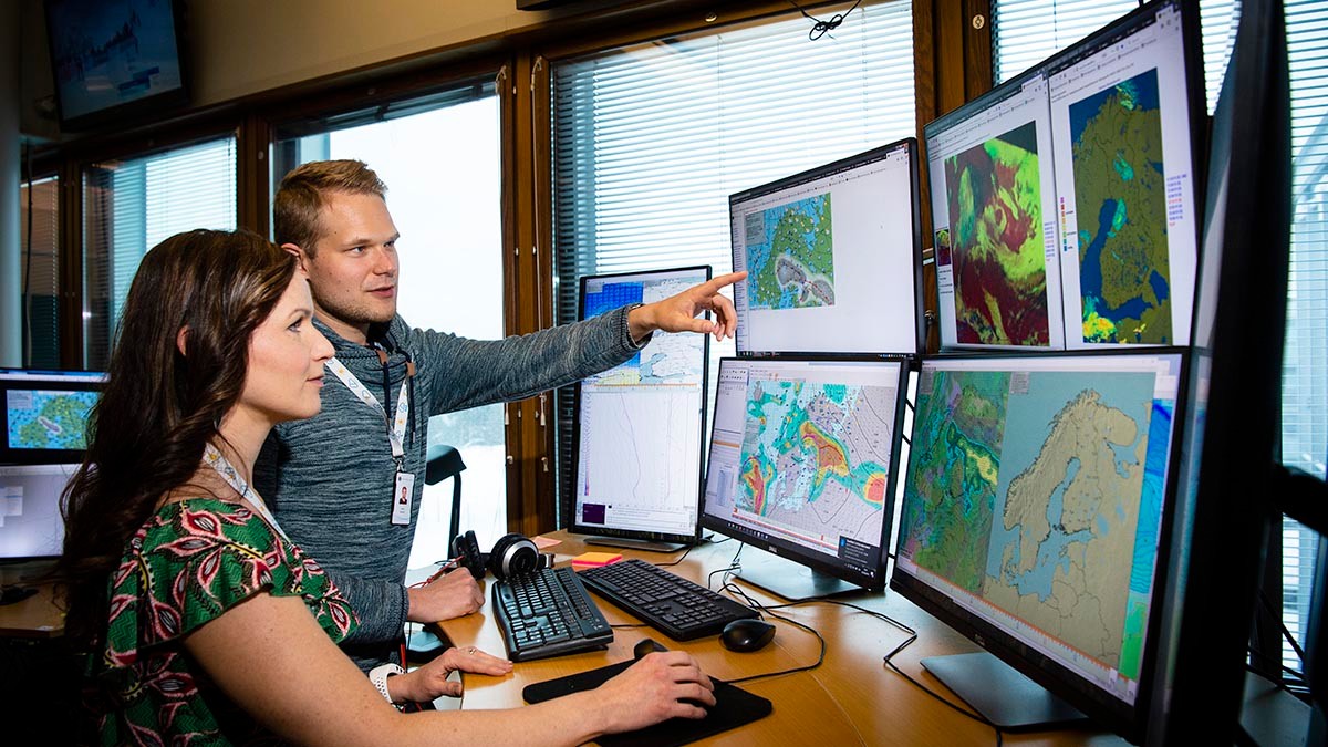 Two meteorologists forecasting the weather. Several monitors in front.